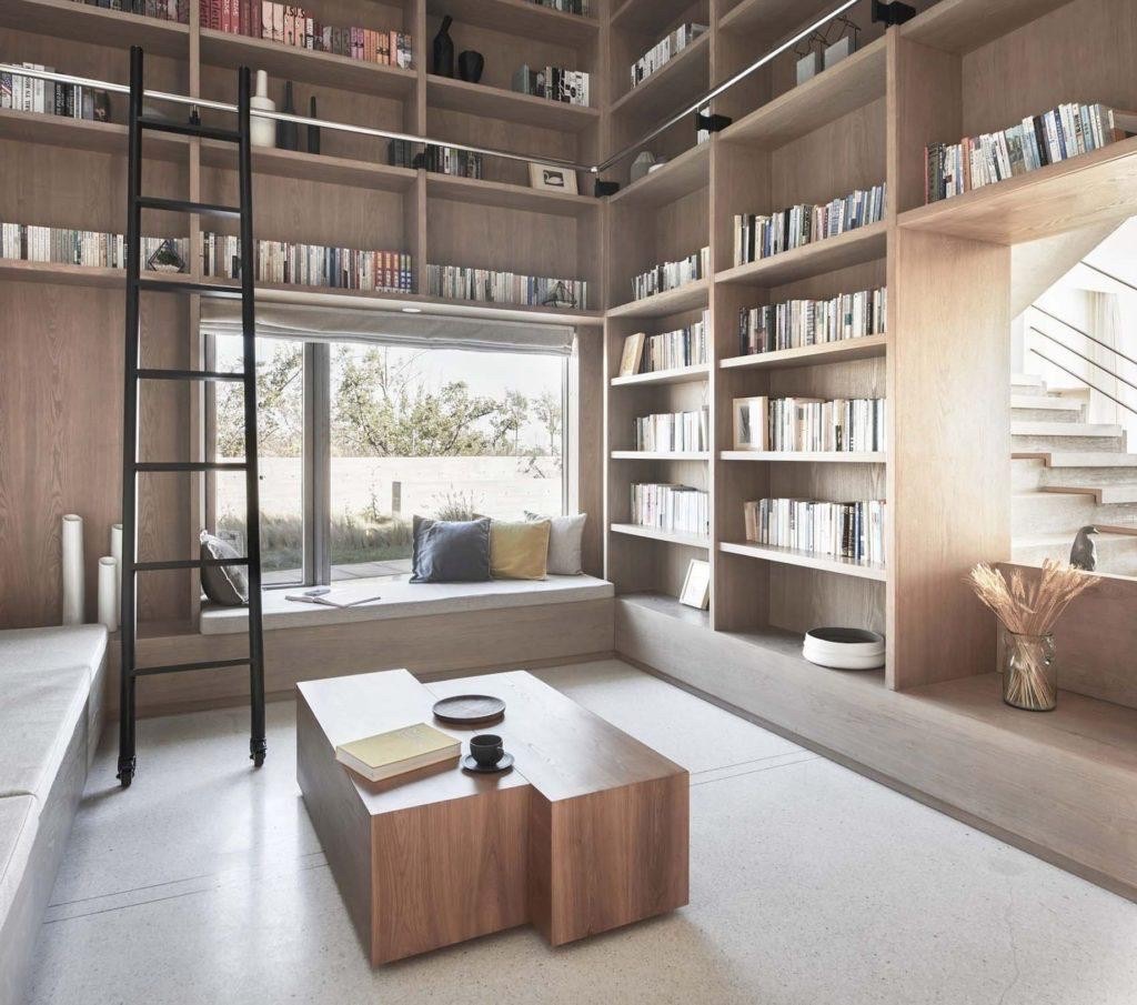 Modern library with floor-to-ceiling wooden bookshelves, a rolling ladder, and a window seat in natural light